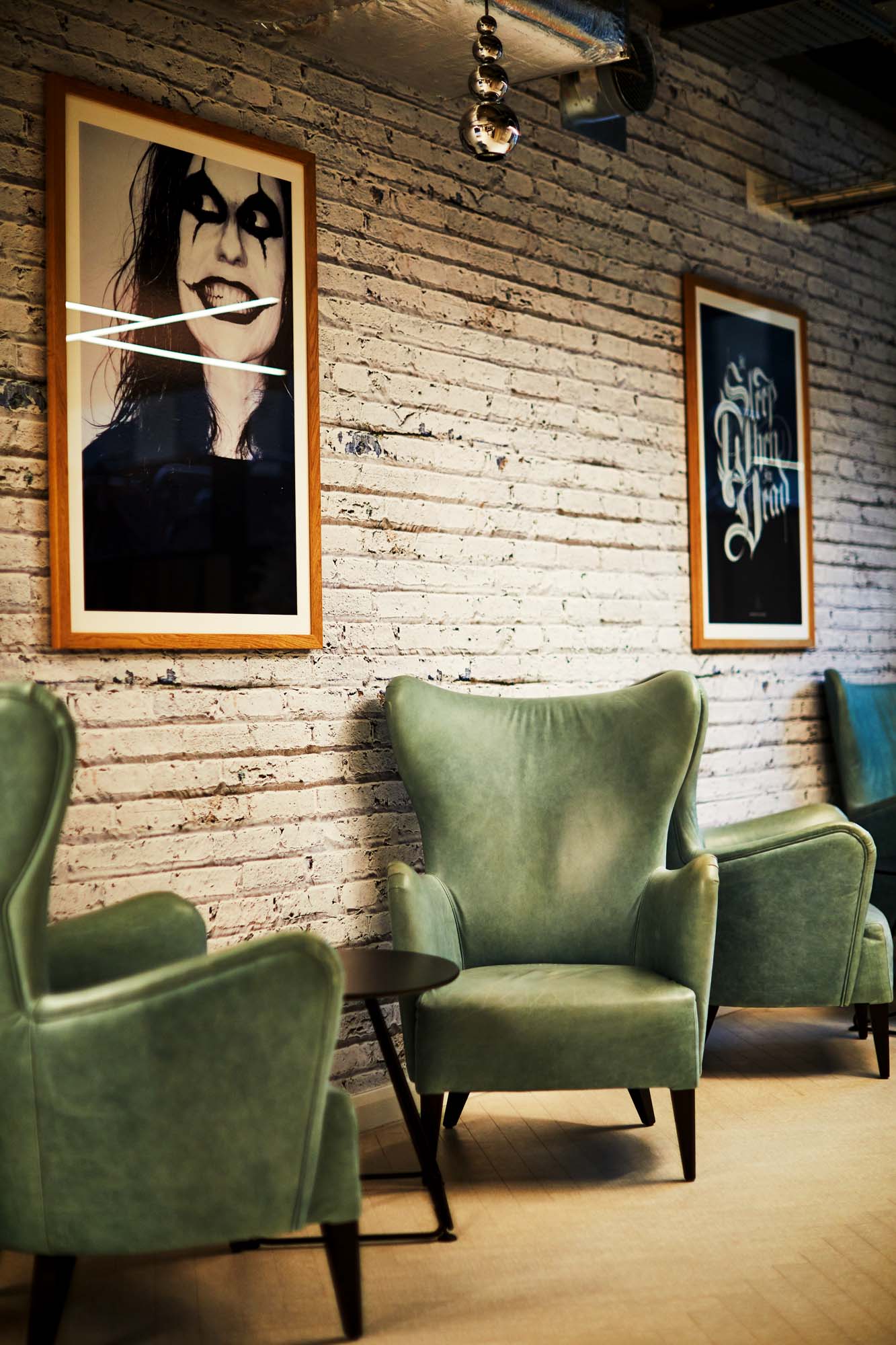 A modern lounge area with two green armchairs and a small circular table. The wall is exposed white brick, featuring two framed artworks: one of a face with dramatic makeup and another with bold text. The floor is light wood.